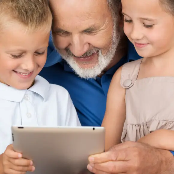 Ein älterer Herr surft mit einem Tablet im Internet umgeben von seinen Enkelkindern.