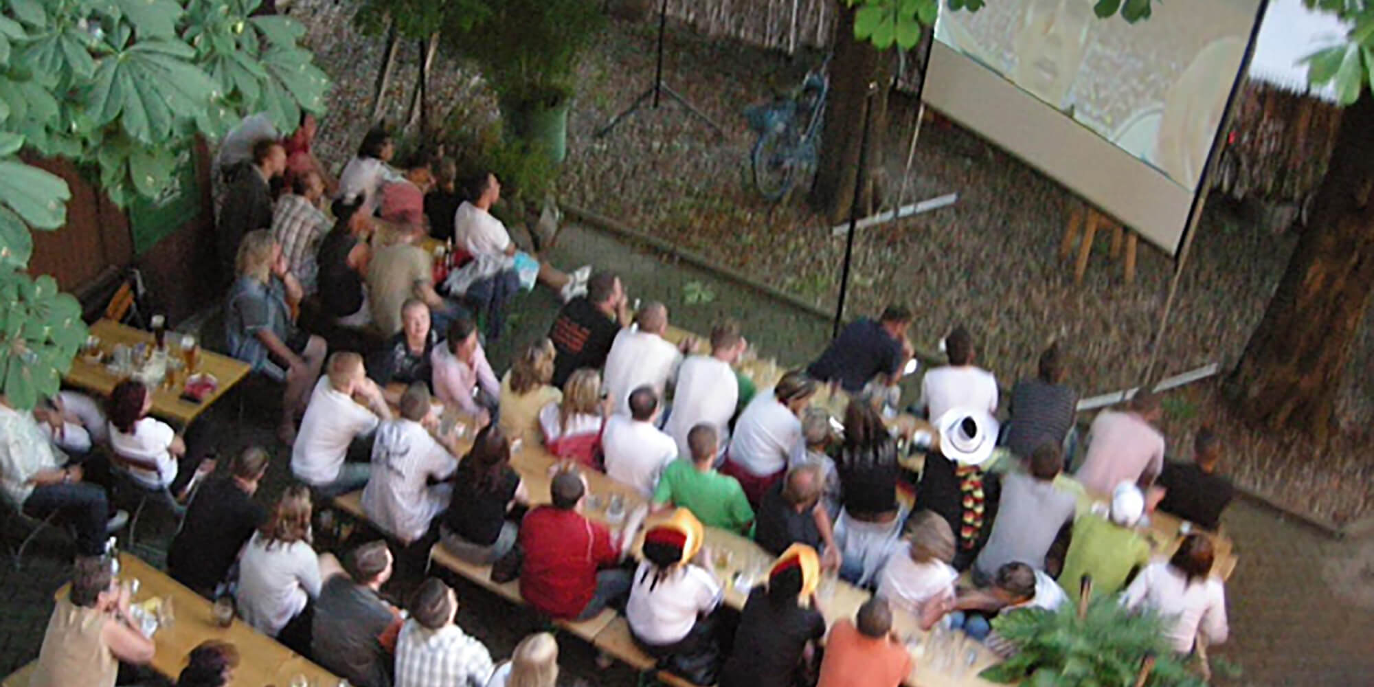 Große Personengruppe schaut Fußball auf Leinwand im Biergarten.